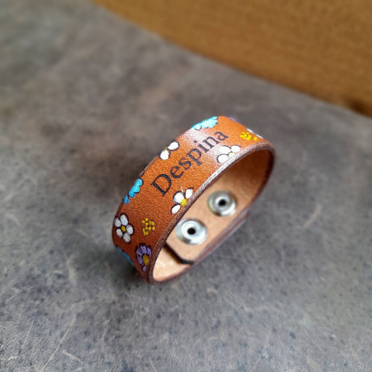 The Bloom & Name Bracelet, a hand-painted floral leather cuff. Shows a custom engraved name on the front, surrounded by small hand-painted flowers, and stainless steel snap buttons.
