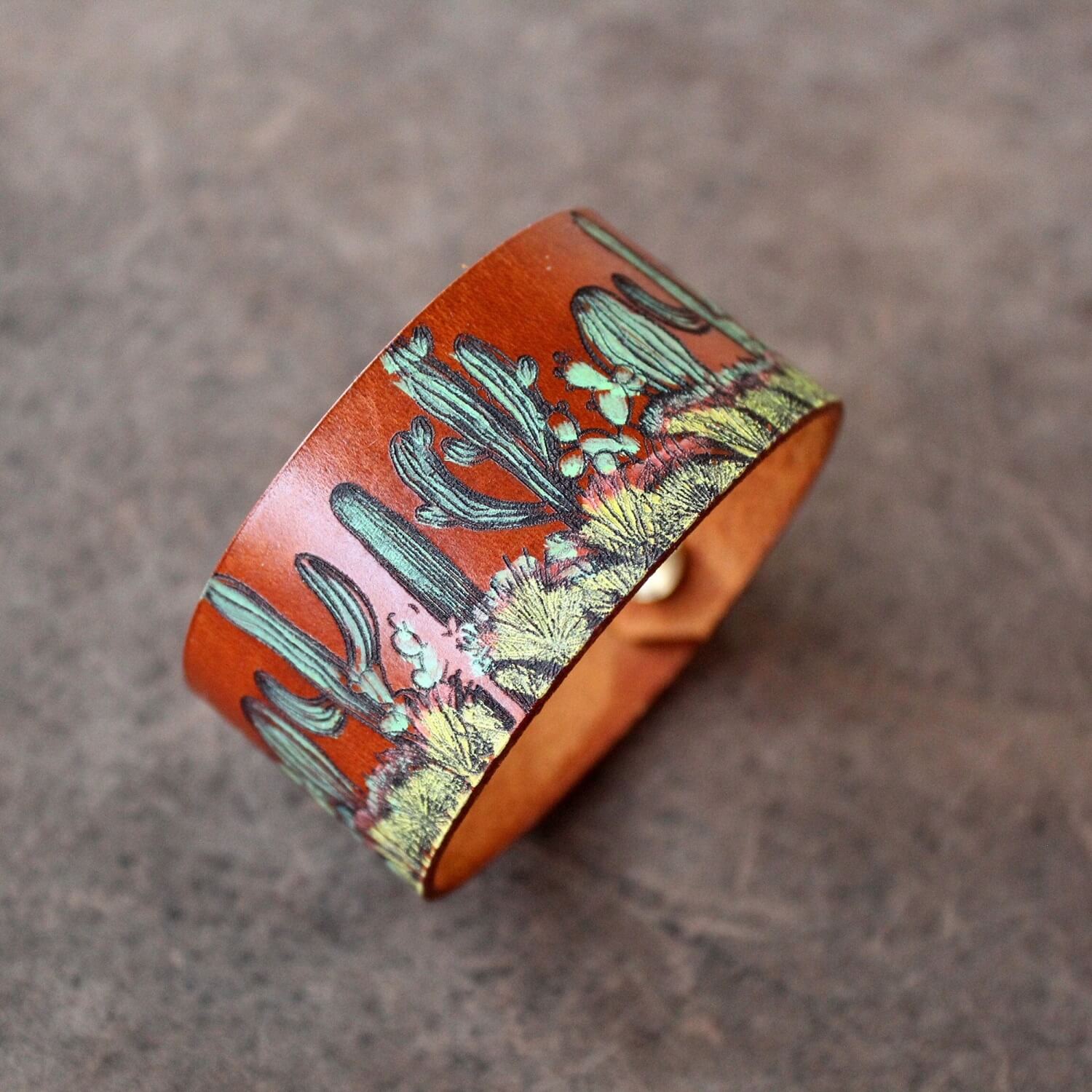 The Desert Bloom Cuff, a hand-painted leather bracelet featuring a vibrant cactus design. Shows the wide leather band and visible snap buttons.