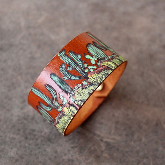 The Desert Bloom Cuff, a hand-painted leather bracelet featuring a vibrant cactus design. Shows the wide leather band and visible snap buttons.