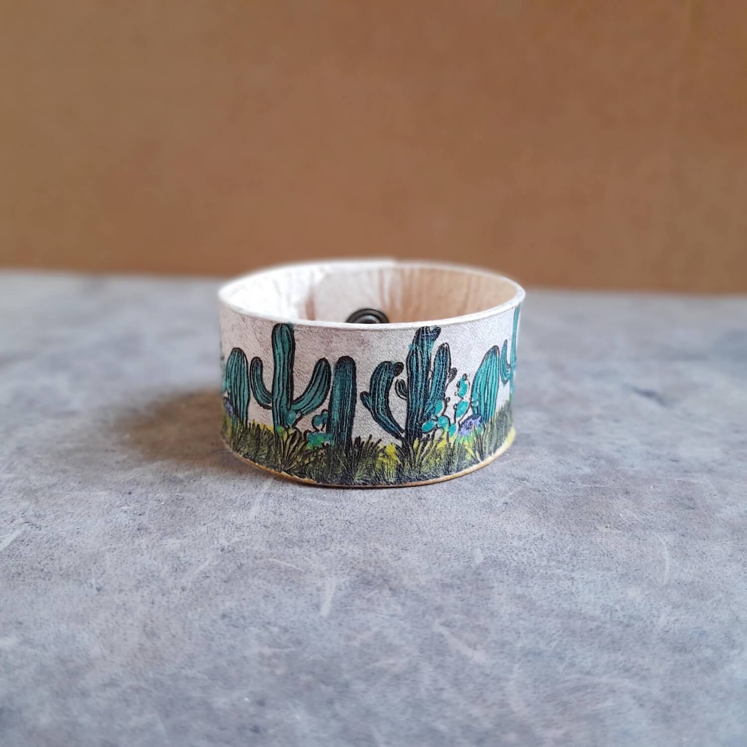 The Desert Bloom Cuff, a hand-painted leather bracelet featuring a vibrant cactus design. Shows the wide leather band and visible snap buttons, in distressed white variation.