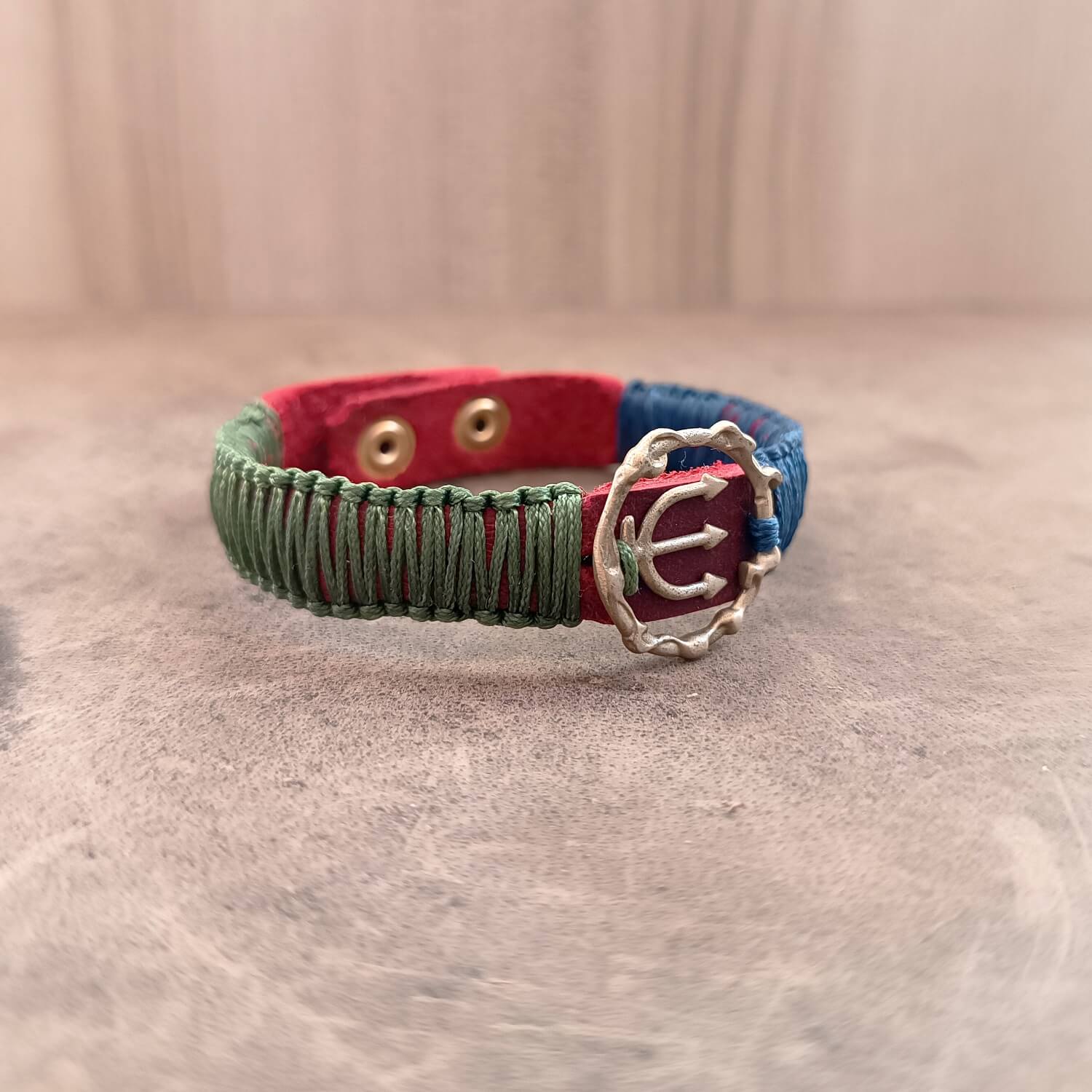 The Guardian's Balance Trident Bracelet from the Soulful Echoes Collection, laid flat. It features a striking brass trident pendant on a vibrant red Crazy Horse leather band, with deep forest green and navy blue macrame cords.