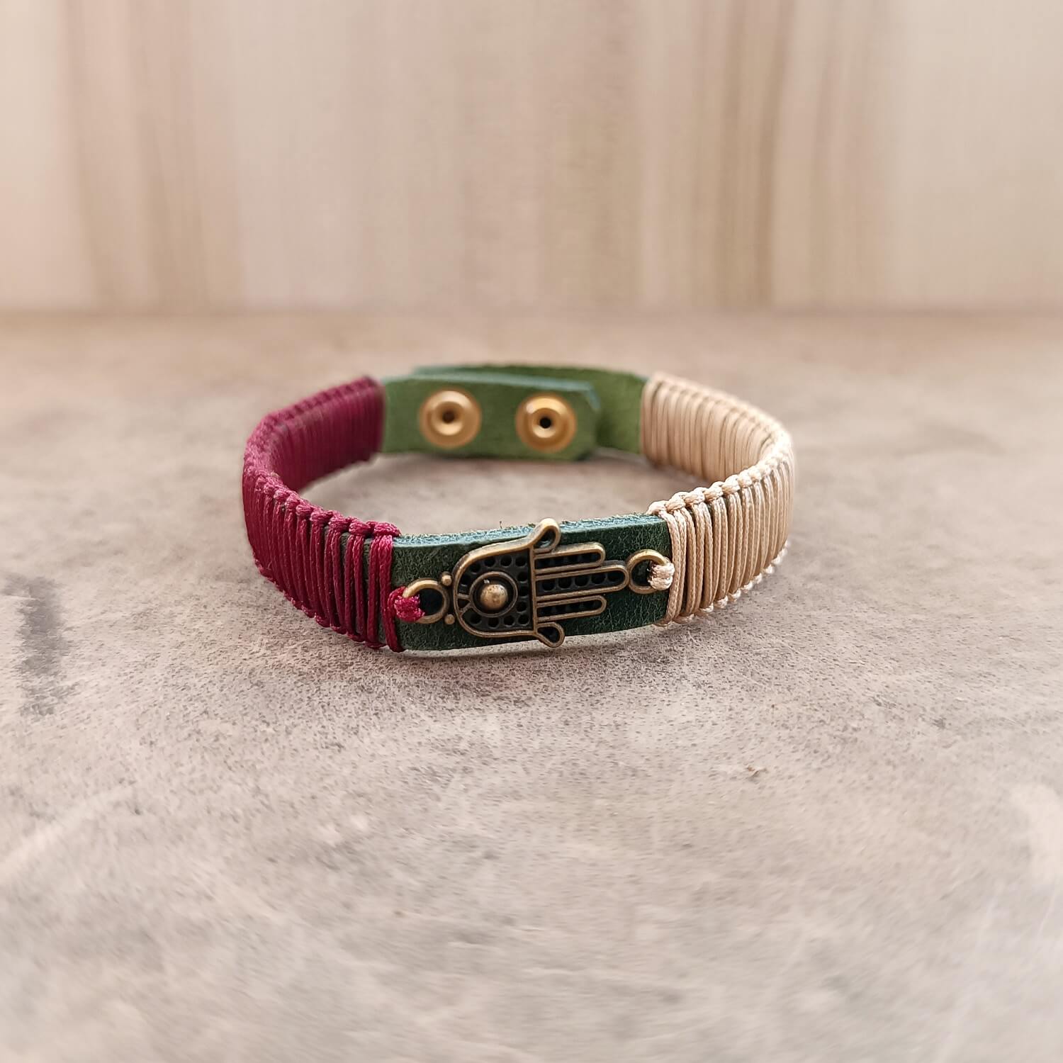 The Hamsa Harmony Bracelet from the Soulful Echoes Collection, laid flat. It features an antique bronze Hamsa Hand pendant on a khaki Crazy Horse leather band, with light bronze and burgundy red macrame cords.