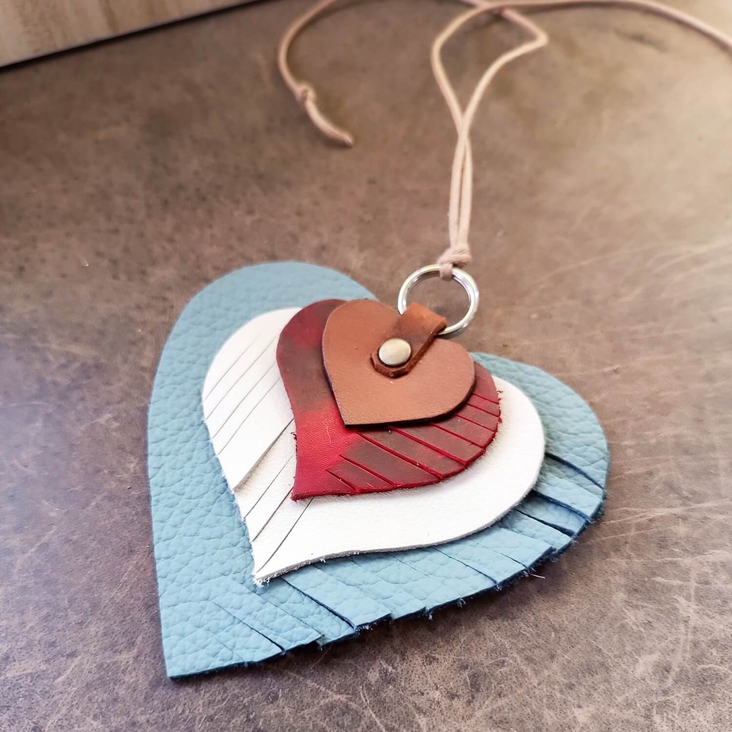 The "Heartstrings" Charm laid flat: a unique, four-layered heart ornament crafted from upcycled leather remnants in brown, burgundy red, white, and light blue, with rotating fringe patterns and a cotton cord.
