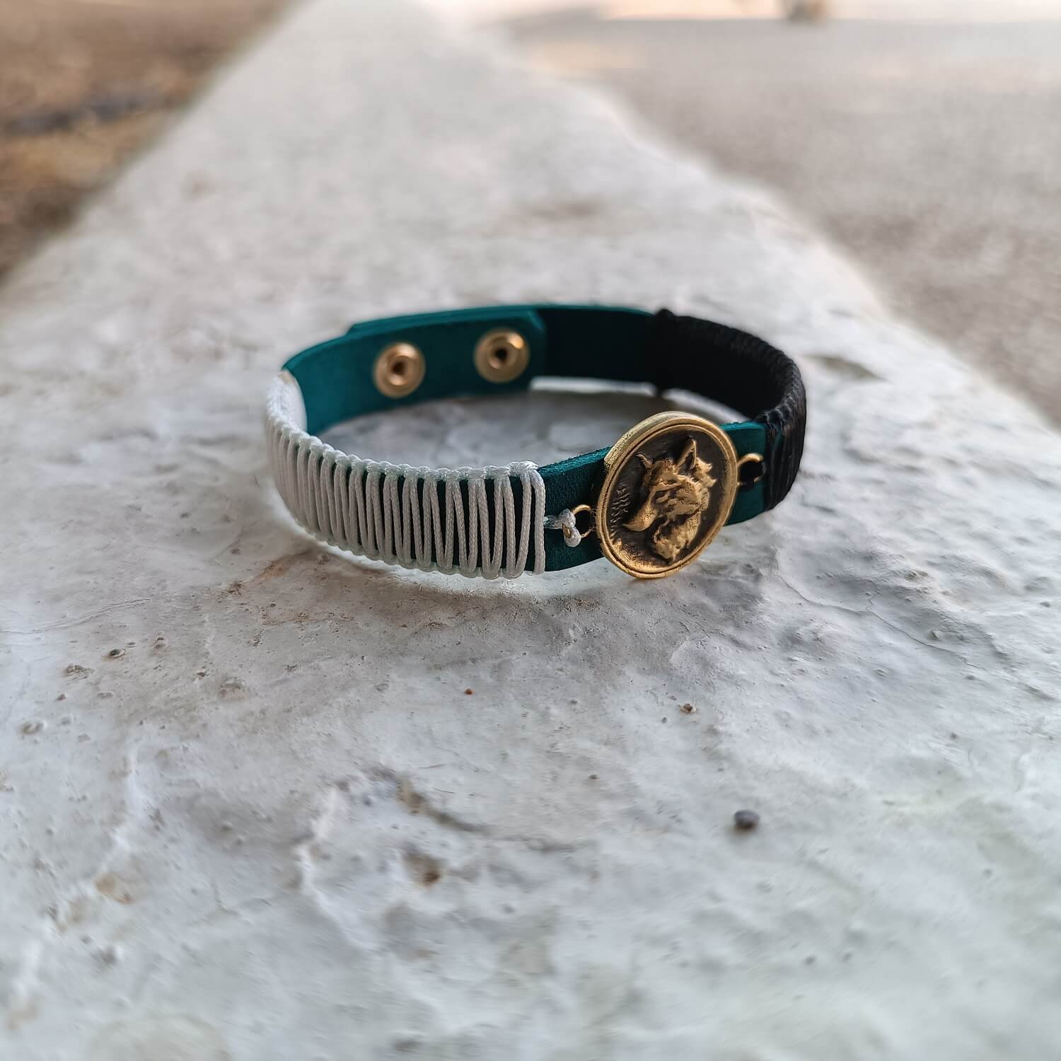 The Instinctive Shield Wolf Bracelet from the Soulful Echoes Collection, laid on urban stepping stone in Athens. It features a detailed oxidized brass wolf pendant on a blue Crazy Horse leather band, with white and black macrame cords.