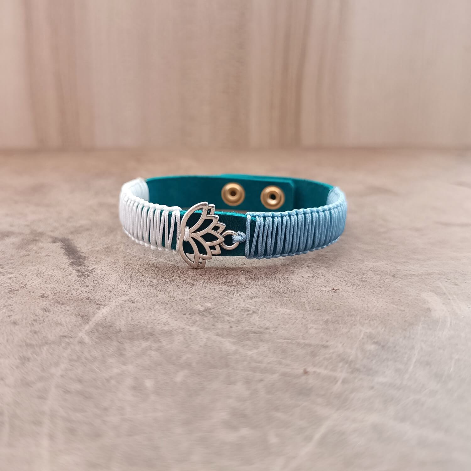 The Lotus's Serenity Bracelet from the Soulful Echoes Collection, laid flat. It features a polished stainless steel lotus flower pendant on a blue Crazy Horse leather band, with white and light blue macrame cords.