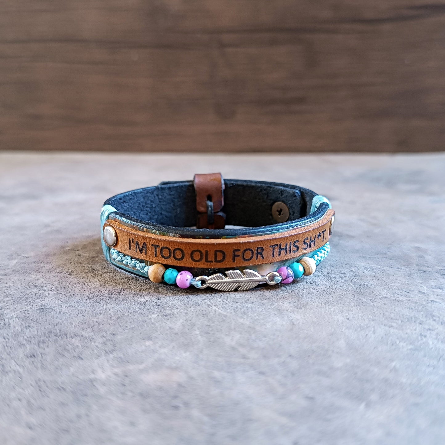 The "No BS" Leather Wrap Bracelet. Features a black band hand-dyed with abstract colorful stripes, a thin leather strip laser-engraved with "I'm Too Old for This Sh*t," a row of colorful beads, and a silver-tone feather charm.