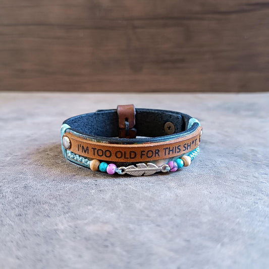 Close up front view of the "No BS" Leather Wrap Bracelet. Features a black band hand-dyed with abstract colorful stripes, a thin leather strip laser-engraved with "I'm Too Old for This Sh*t," a row of colorful beads, and a silver-tone feather charm.