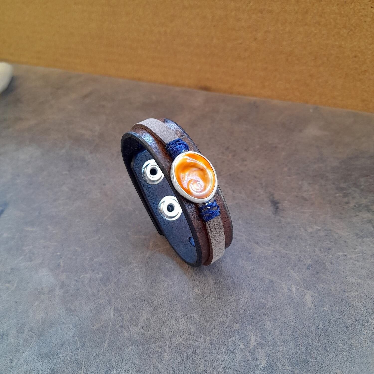 The OOAK 'Eye of the Sea' Bracelet: A layered dark brown and cigar gray full-grain leather band featuring a natural Eye of Saint Lucia sea shell set on a sterling silver 925 pendant, wrapped with deep navy blue waxed cord.