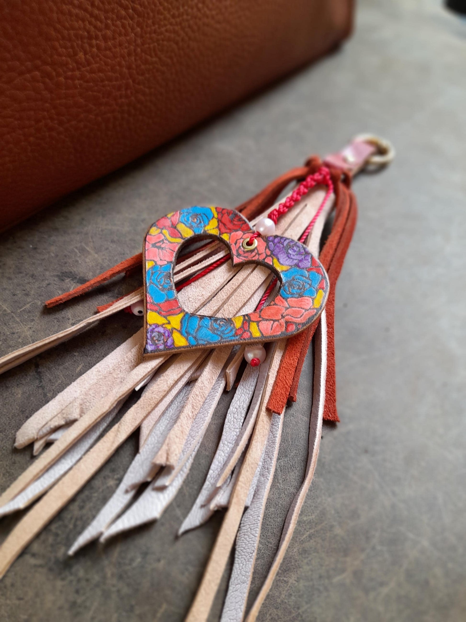 The OOAK Stained Glass Heart Charm: A hand-painted floral leather "hollow heart" with vibrant colors, attached to a flowing pink suede tassel with red fringe. Features a gold-tone O-ring clip.