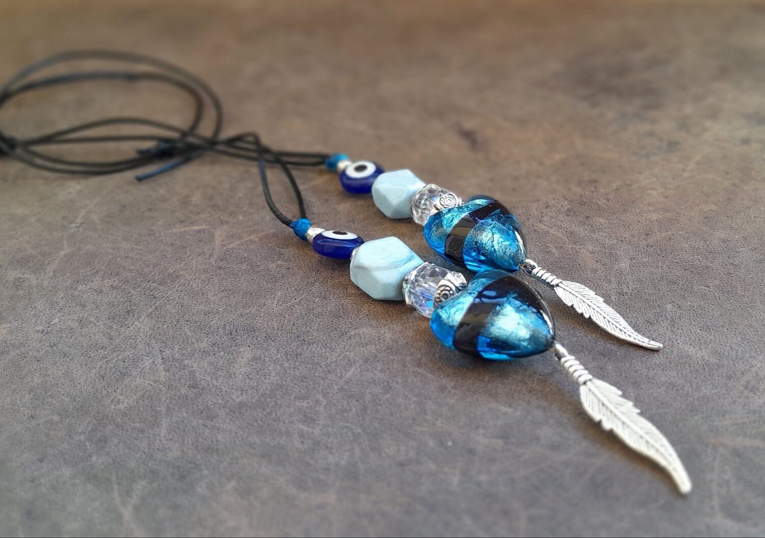 A turquoise and silver car charm with a black cord, featuring a large heart-shaped turquoise stone, a silver feather, and a small evil eye detail.