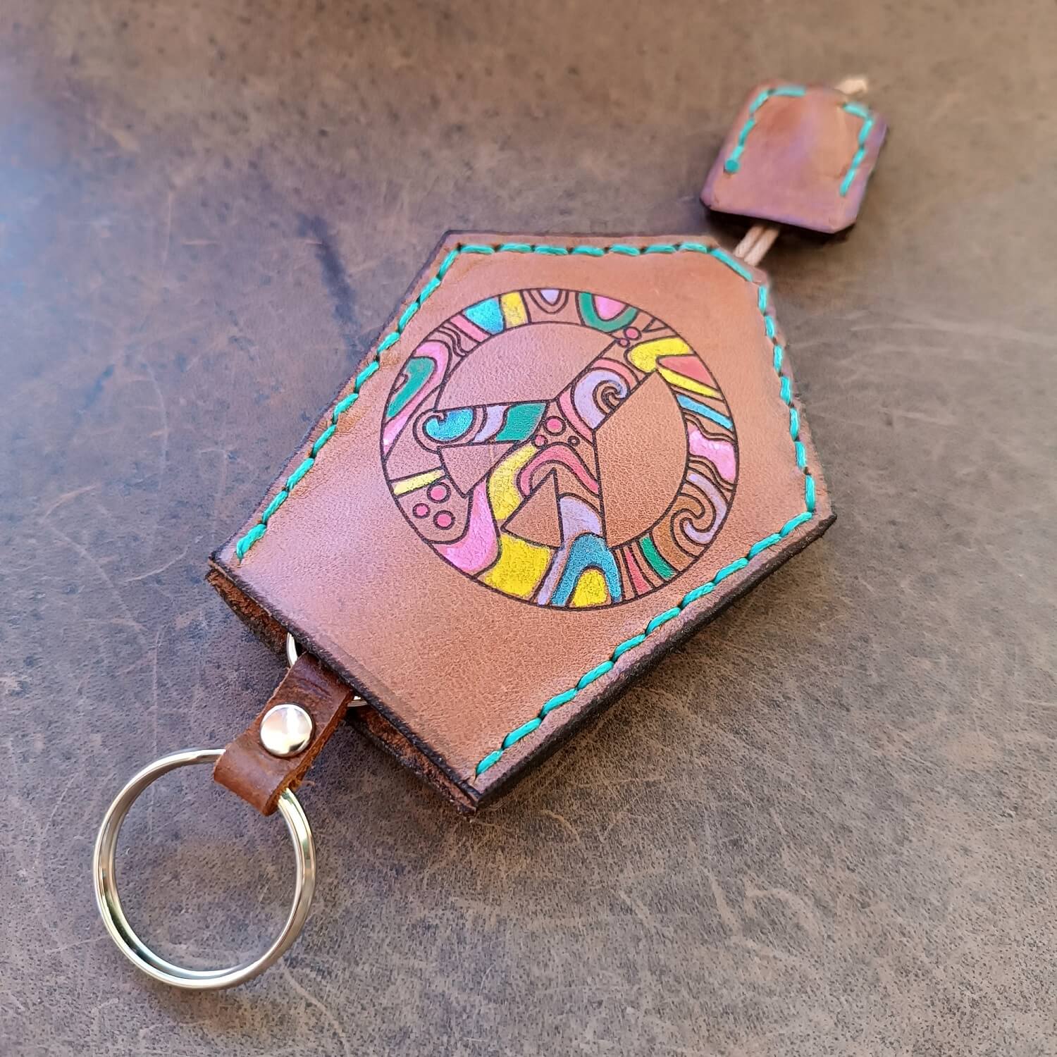 The Peace Weaver Boho Leather Key Holder. Made from brown Vachetta leather with a hand-painted, laser-engraved peace symbol in colorful abstract patterns, and vibrant teal/turquoise stitching. Shows the pull strap for keys.