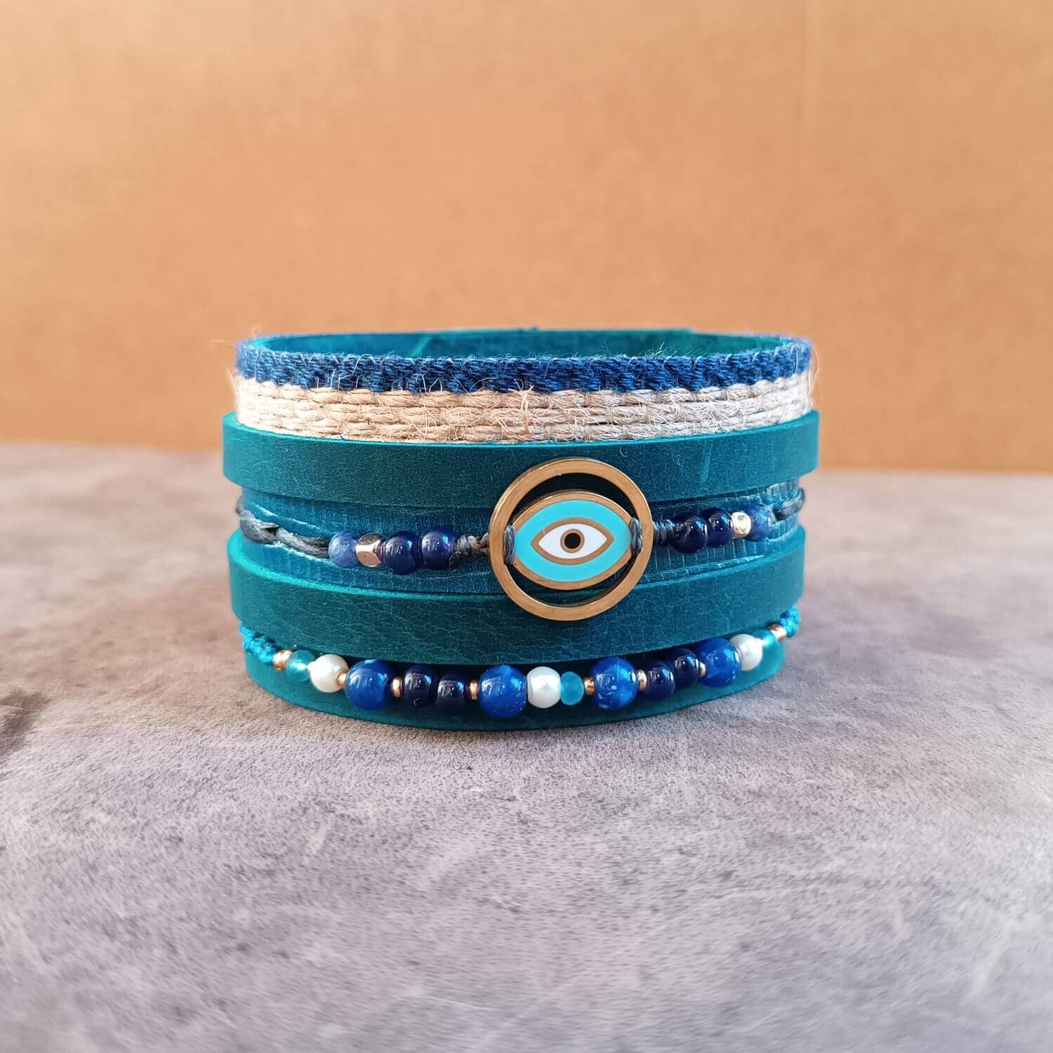close-up of the gold-tone Evil Eye pendant on The Protector Cuff, surrounded by sodalite, lapis lazuli, turquoise, and white howlite gemstone beads on macramé cords.