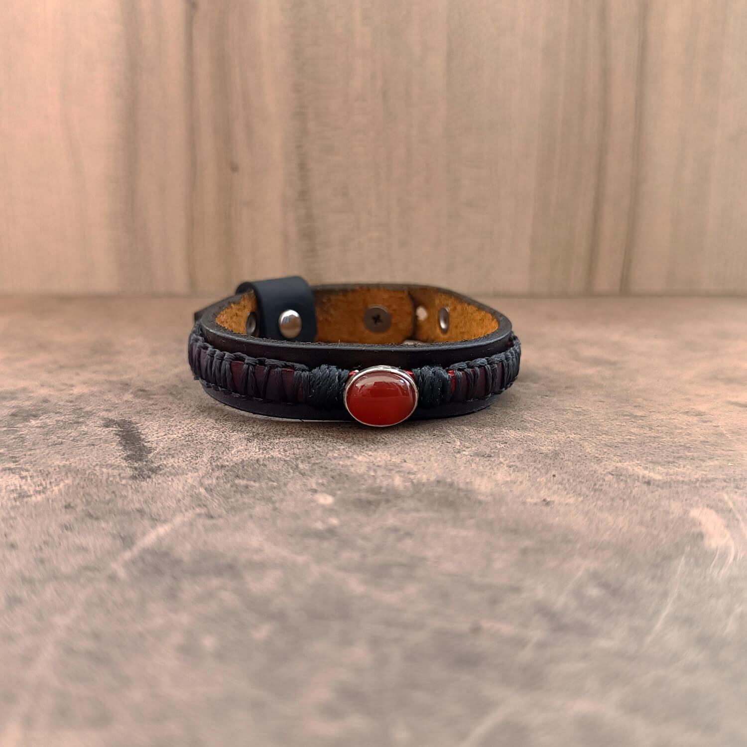 The Roamer Leather Bracelet, a men's layered leather bracelet, laid flat. Shows the dark brown/black bottom band, braided crimson red leather, and a sterling silver pendant with a red Carnelian gemstone at its center.