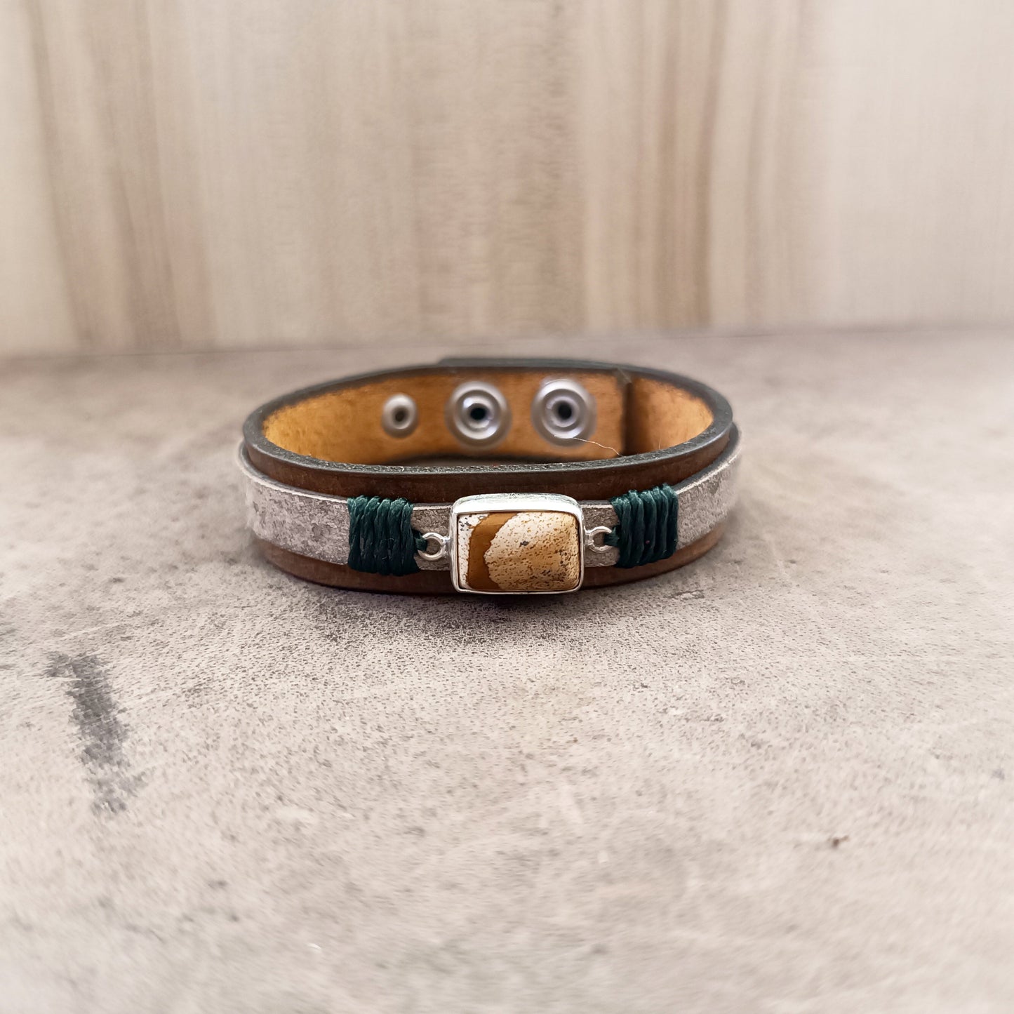 The Silent Witness Bracelet, a one-of-a-kind men's leather bracelet, laid flat. Features a natural Picture Jade stone set in sterling silver, layered over hand-dyed dark brown and marbled beige vachetta leather with forest green cord accents.