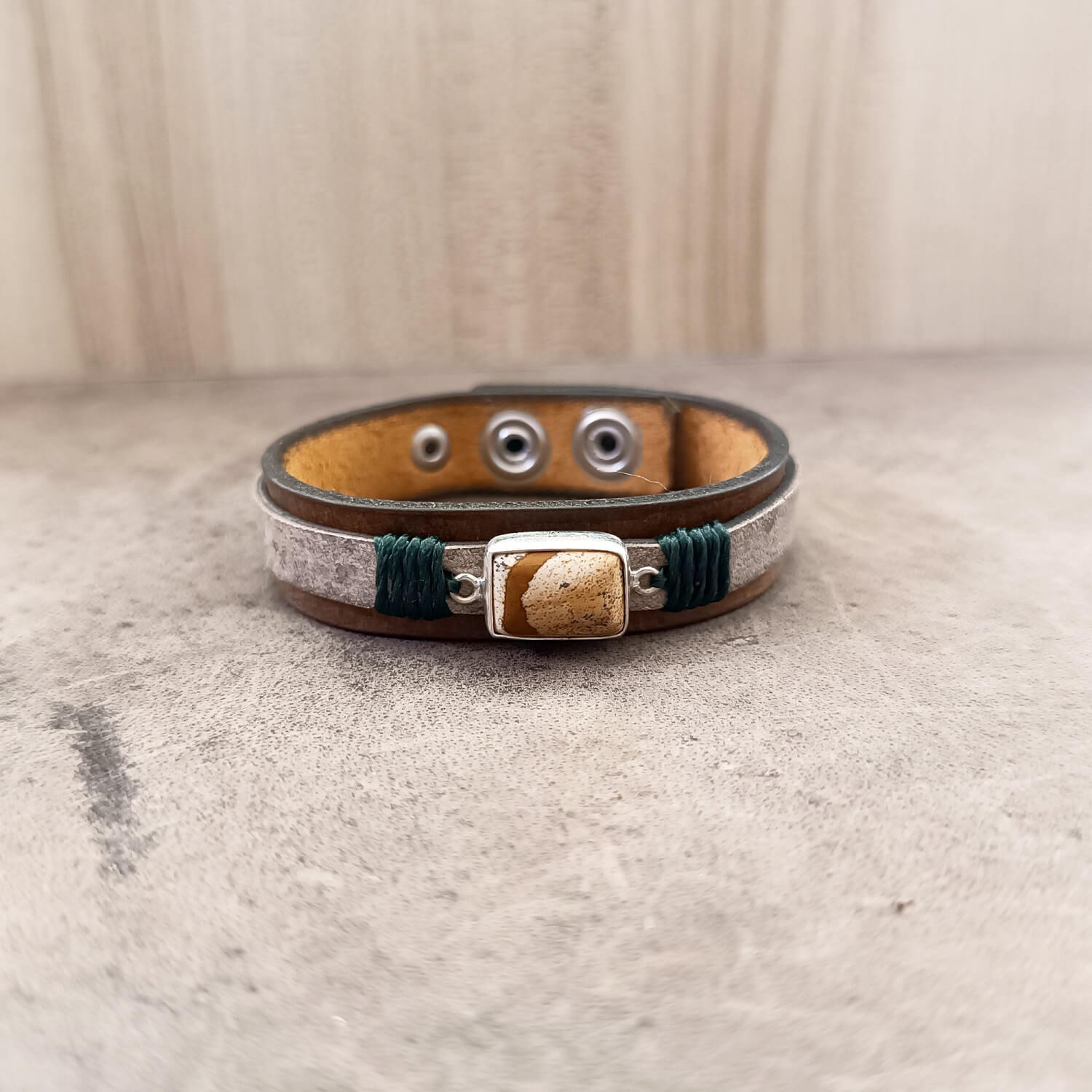 The Silent Witness Bracelet, a one-of-a-kind men's leather bracelet, laid flat. Features a natural Picture Jade stone set in sterling silver, layered over hand-dyed dark brown and marbled beige vachetta leather with forest green cord accents.