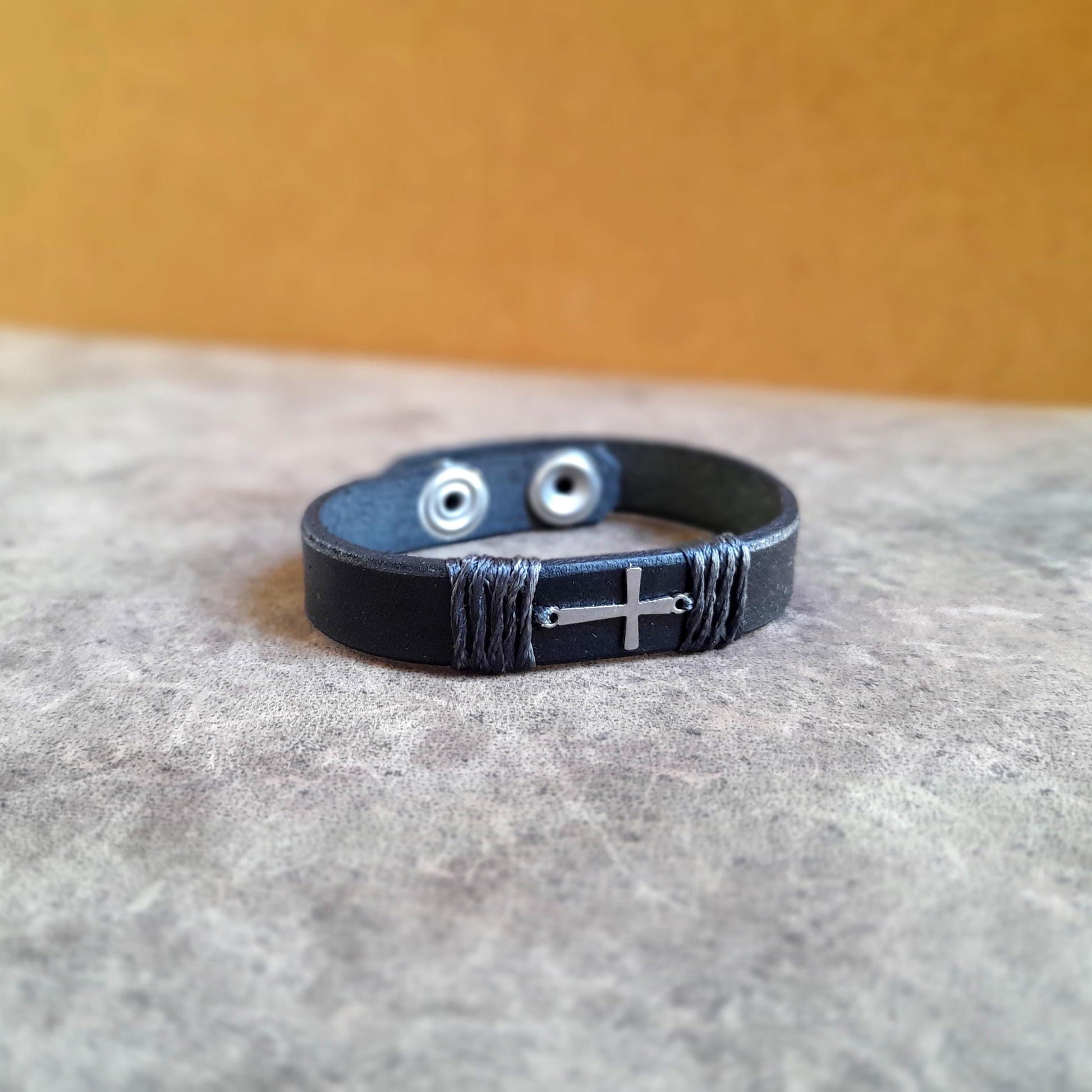 The Subtle Cross Bracelet in Black, a minimalist leather cuff with a tiny horizontal silver stainless steel cross. Shows the thick full grain leather and snap closure.