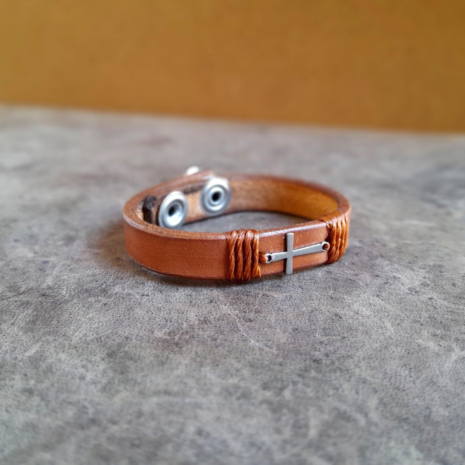 The Subtle Cross Bracelet in Brown, a minimalist leather cuff with a tiny horizontal silver stainless steel cross. Shows the thick full grain leather and snap closure.