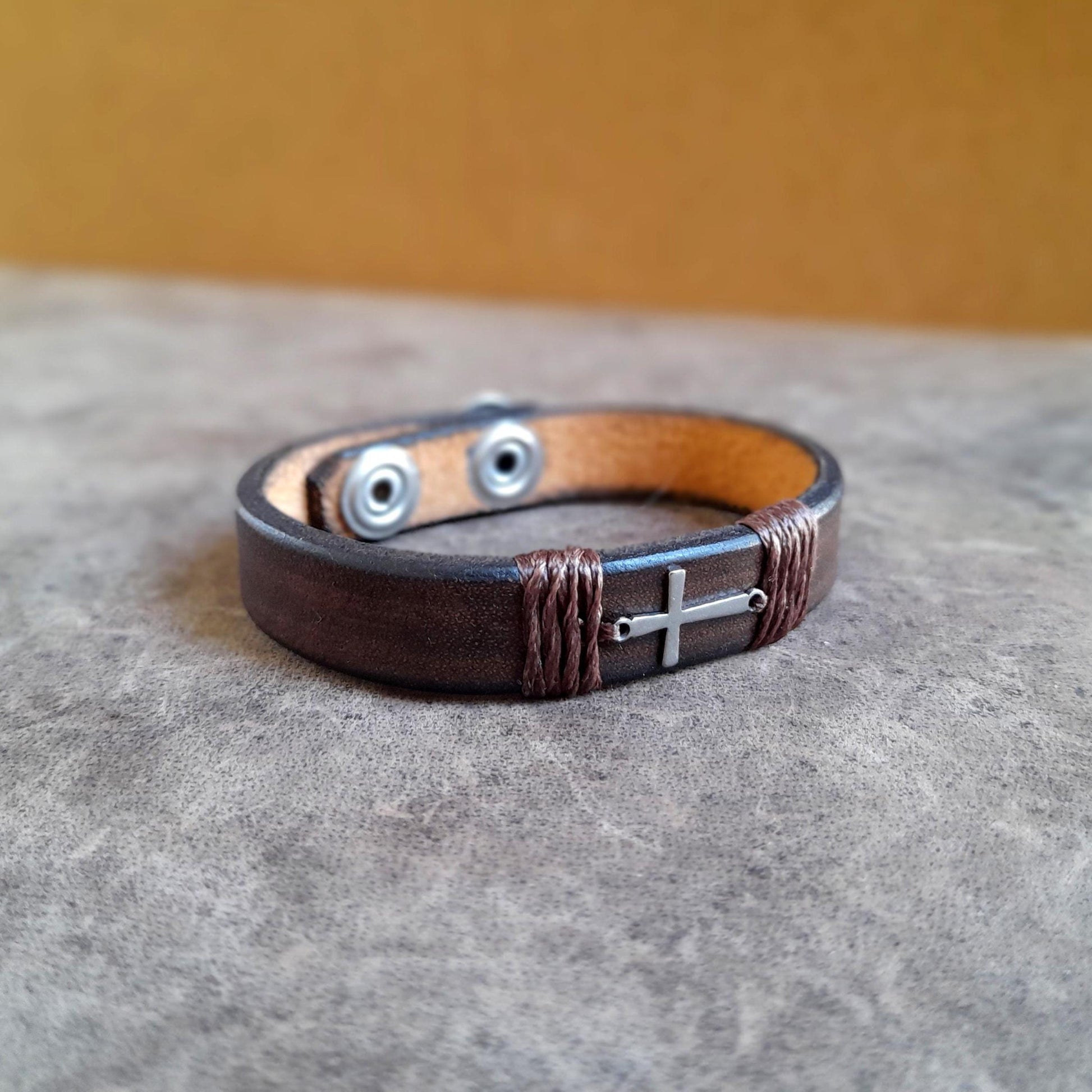 The Subtle Cross Bracelet in Dark Brown, a minimalist leather cuff with a tiny horizontal silver stainless steel cross. Shows the thick full grain leather and snap closure.