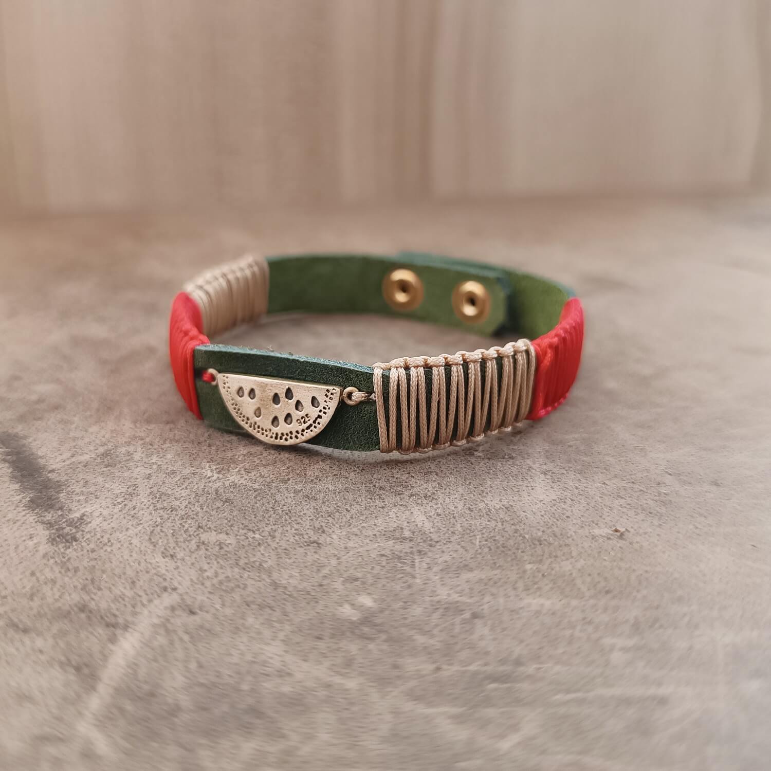 The Sweet Abundance Watermelon Bracelet from the Soulful Echoes Collection, laid flat. It features a charming oxidized sterling silver watermelon slice pendant on a deep forest green Crazy Horse leather band, with vibrant red and light bronze macrame cords.