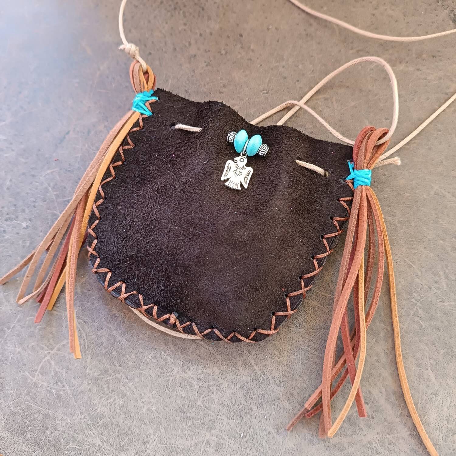 The Thunderbird Keeper Neck Pouch, a limited edition piece, laid flat. Made of chestnut brown suede leather, it features a silver Thunderbird charm and turquoise wooden beads with suede tassels.