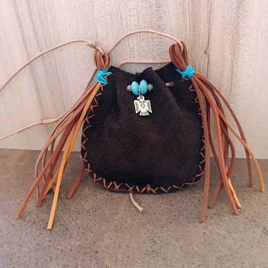 The Thunderbird Keeper Neck Pouch, a limited edition piece. Made of chestnut brown suede leather, it features a silver Thunderbird charm and turquoise wooden beads with suede tassels.