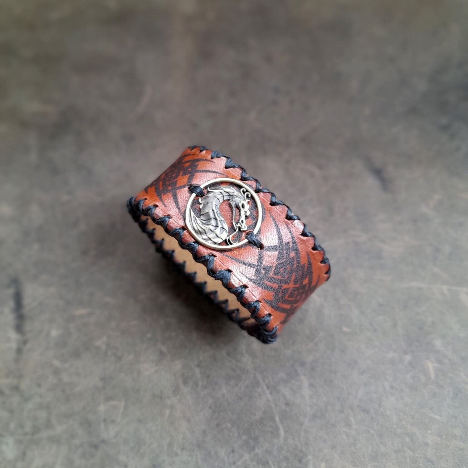 A leather bracelet with a tribal design and a brass dragon pendant, displayed on a textured background.