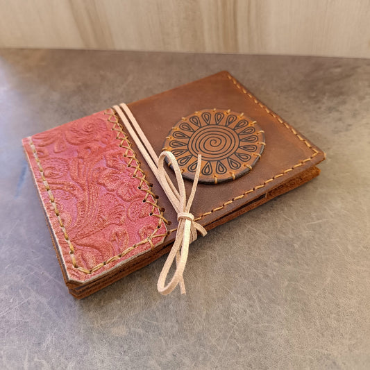 The Wanderer's Journal leather passport case, closed with its soft suede cord, showing the full exterior of the Crazy Horse leather with brick-red floral accents and the spiral sun.
