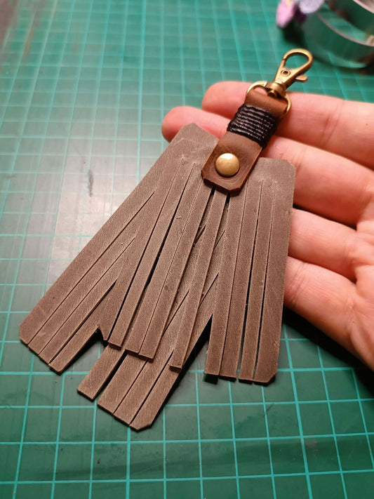 Gray leather fringe keychain held by a hand on a green cutting mat.
