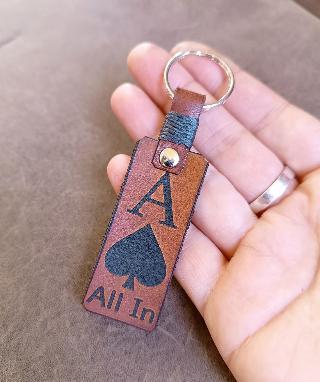 Brown leather keychain with 'All In' and spade symbol held by a hand.