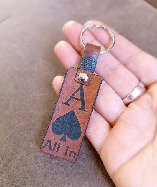 Brown leather keychain with 'All In' and spade symbol held by a hand.
