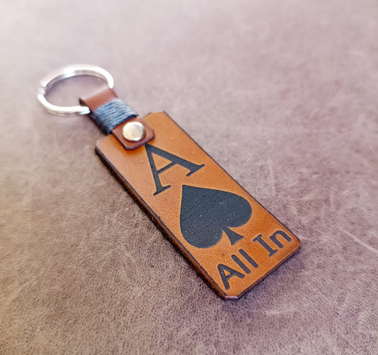 A brown leather keychain with a black ace of spades design and the text 'All In' engraved on it. It features a silver-colored metal ring at the top for attaching keys.