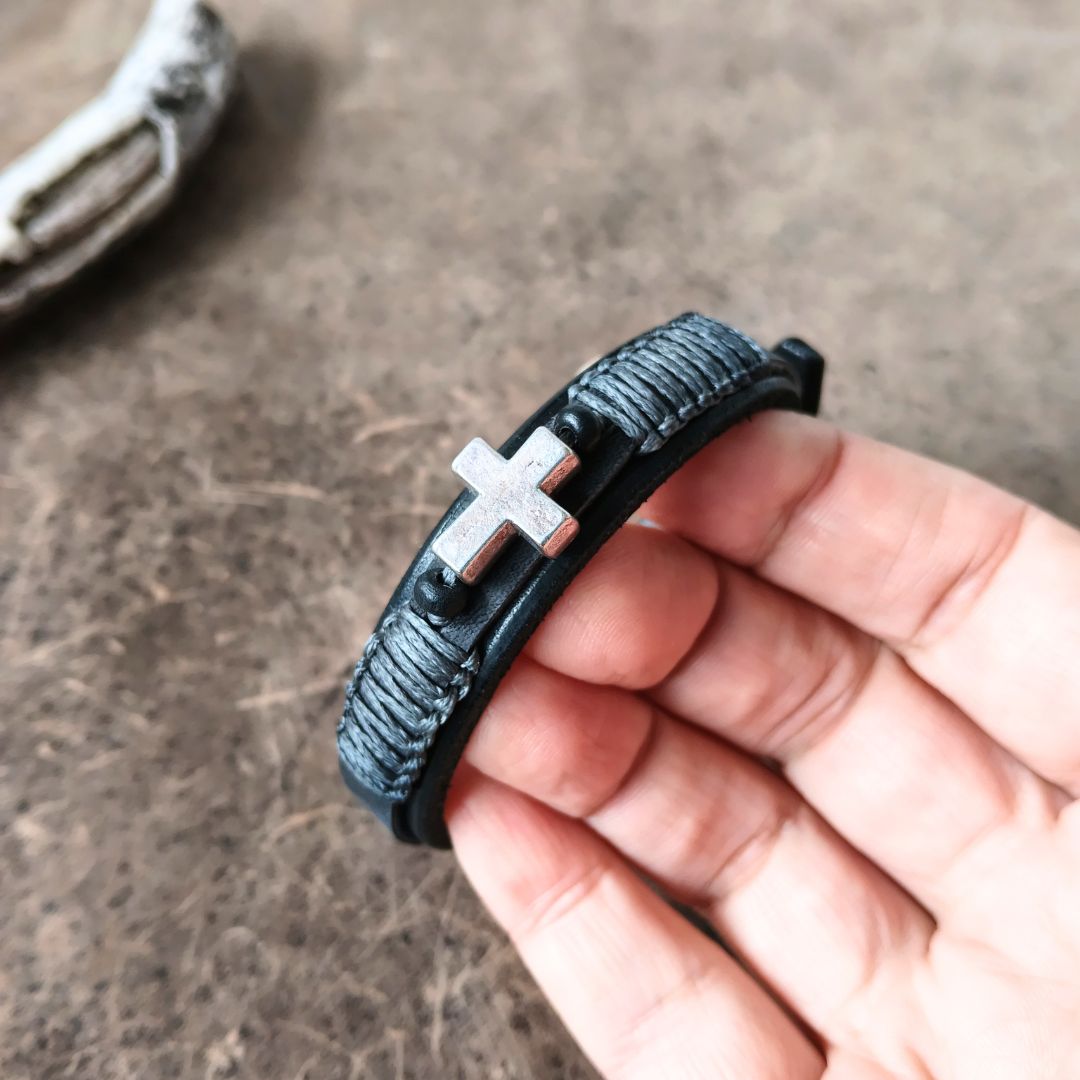 A close-up of a hand holding a black textured leather wrap bracelet with a silver-toned cross charm, set against a rustic brown background.
