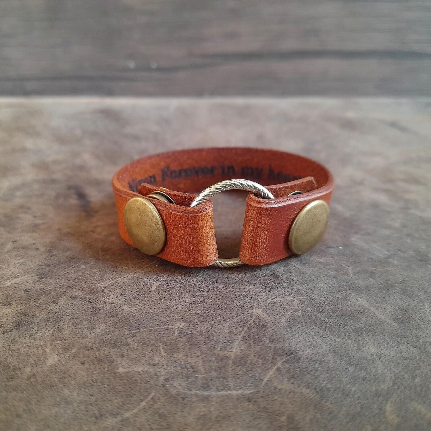 A handmade leather ring holder bracelet in brown, with two metal snap buttons. The bracelet is displayed on a textured background.