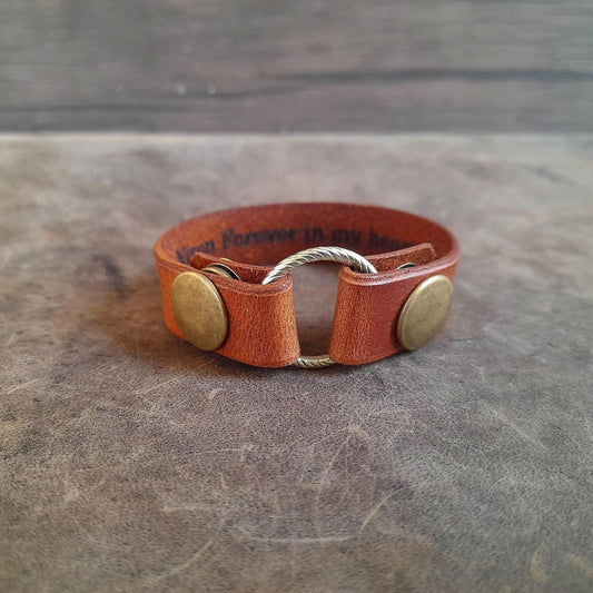 A handmade leather ring holder bracelet in brown, with two metal snap buttons. The bracelet is displayed on a textured background.
