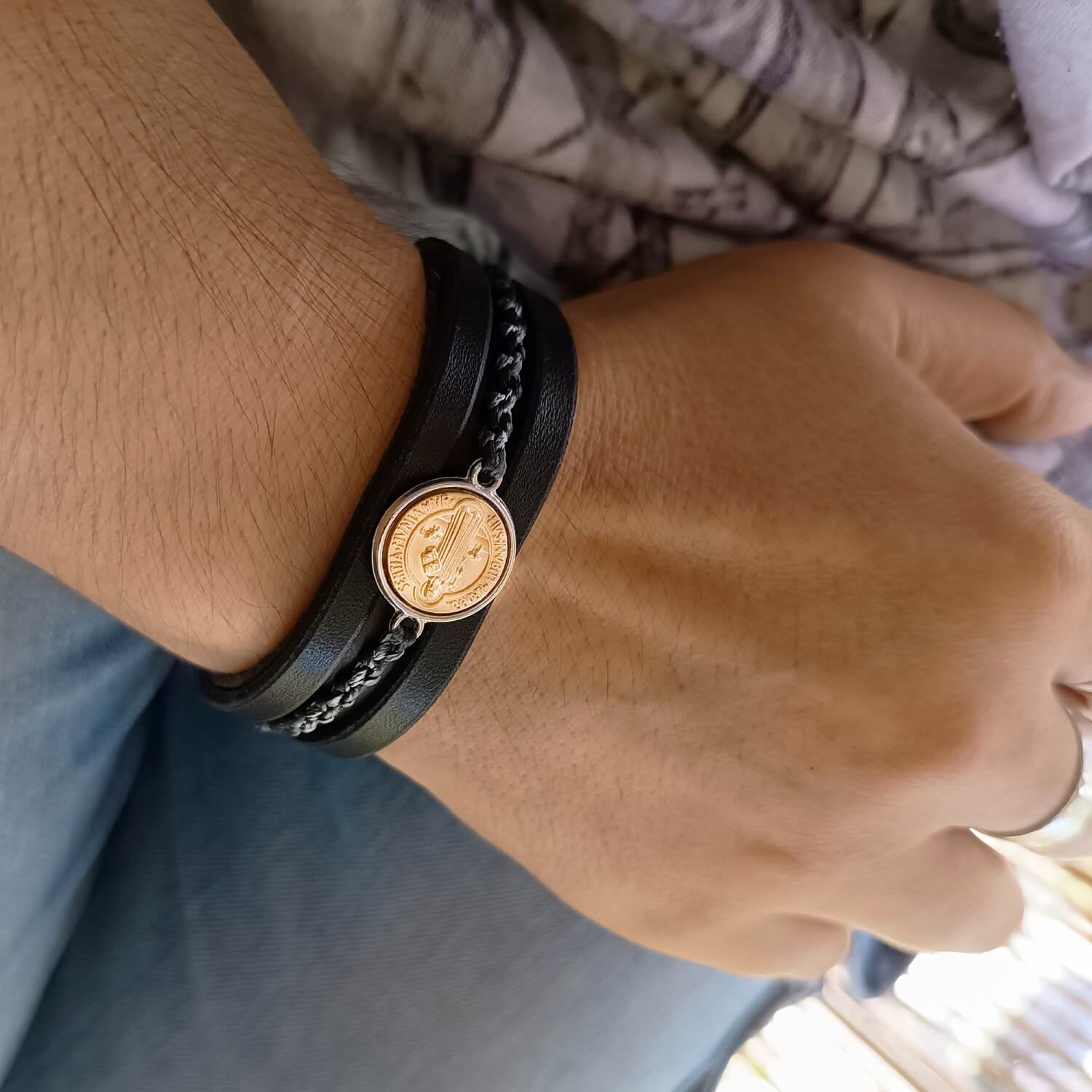 Close-up of a wrist wearing a black leather bracelet with a gold and silver Byzantine style coin pendant.
