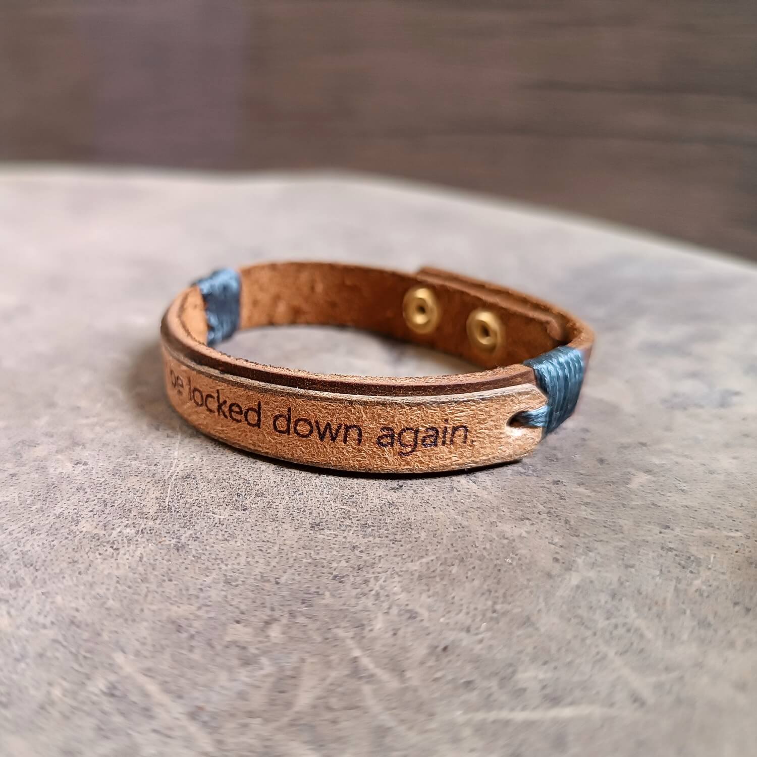 Dark brown and beige leather cuff engraved with “I won’t be locked down again,” adjustable stainless steel buttons, close-up side view showing texture, layers and cord