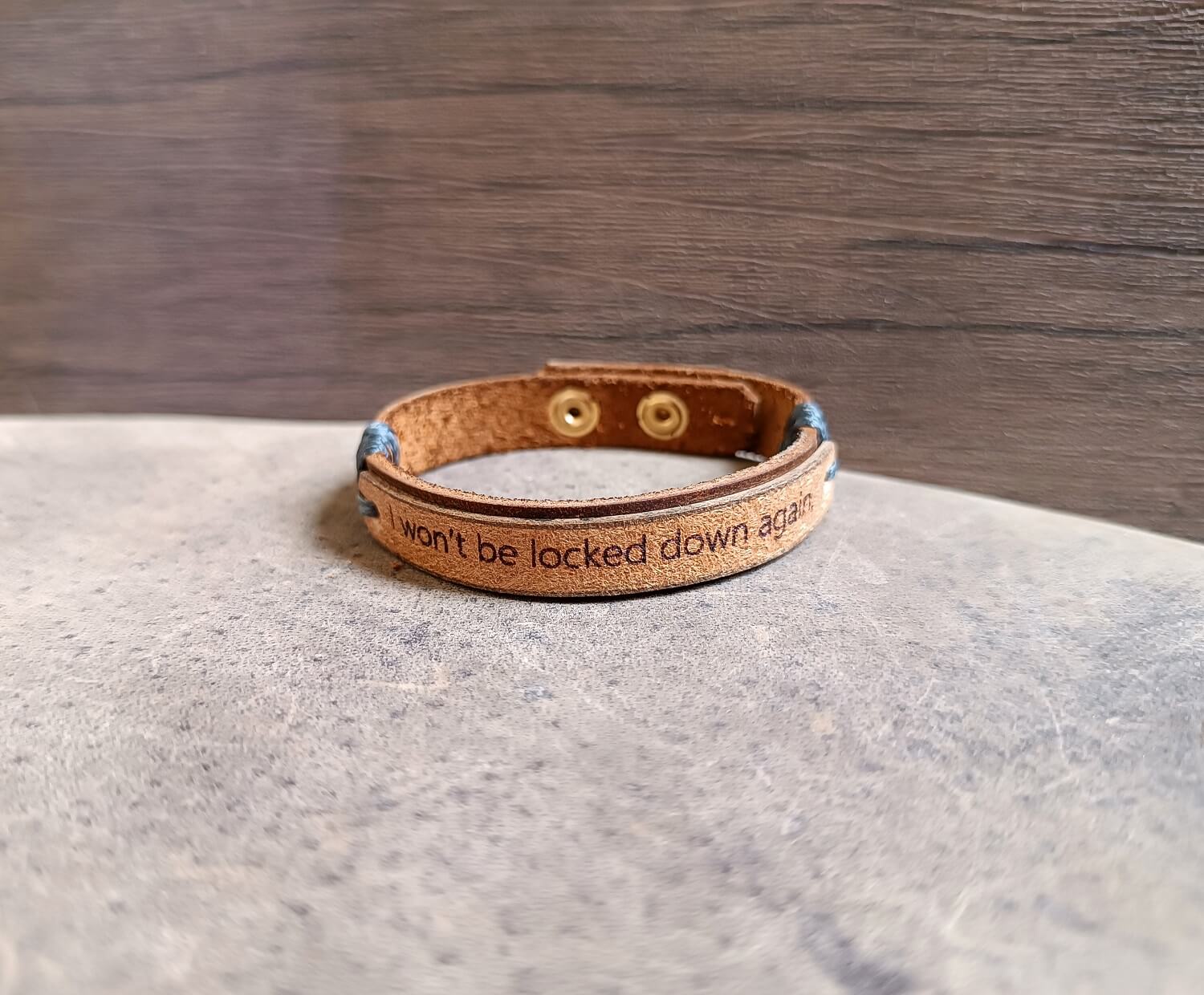 Dark brown and beige leather cuff engraved with “I won’t be locked down again,” adjustable stainless steel buttons, close-up side view showing texture, layers and cord