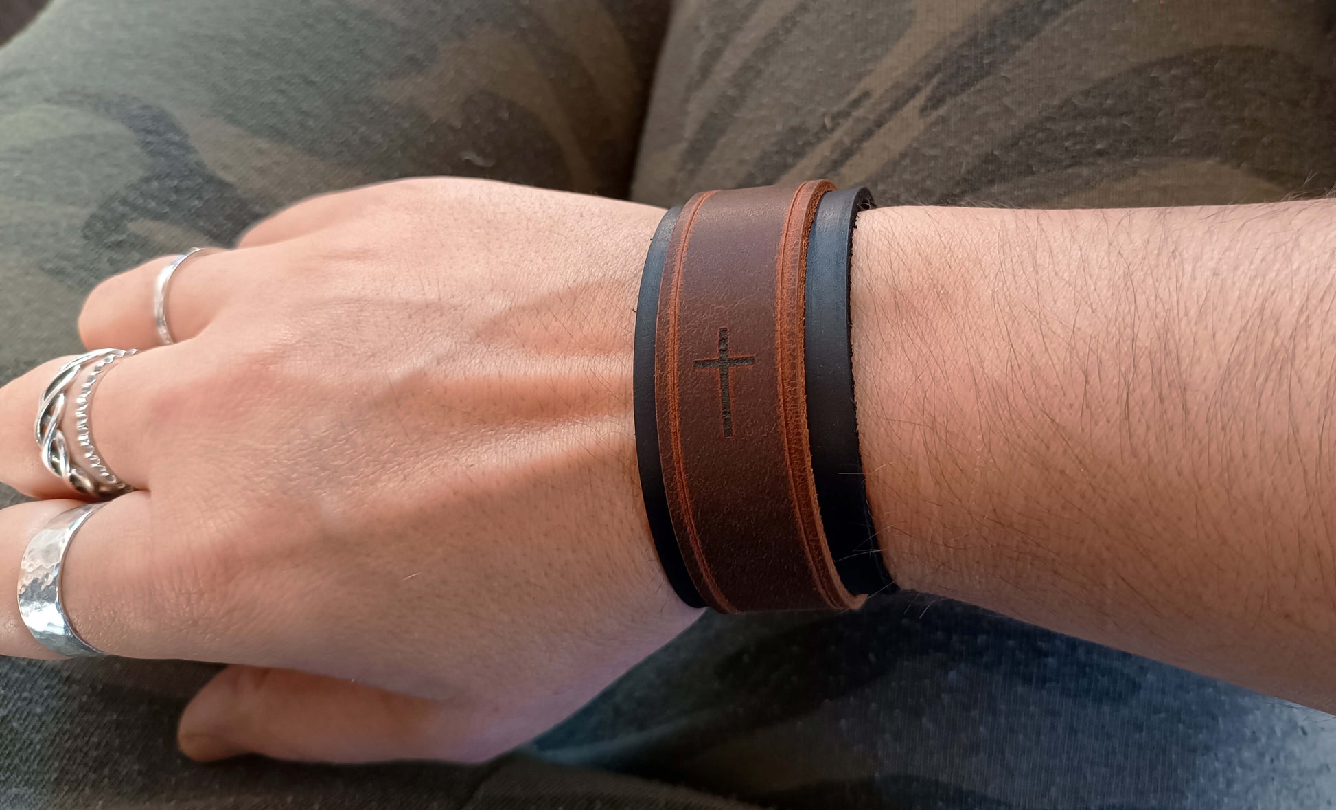 Hand wearing a brown leather bracelet with a cross design on a camouflage-patterned surface.