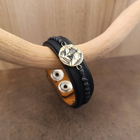 Black brown braided leather bracelet with a brass raven charm on a wooden surface