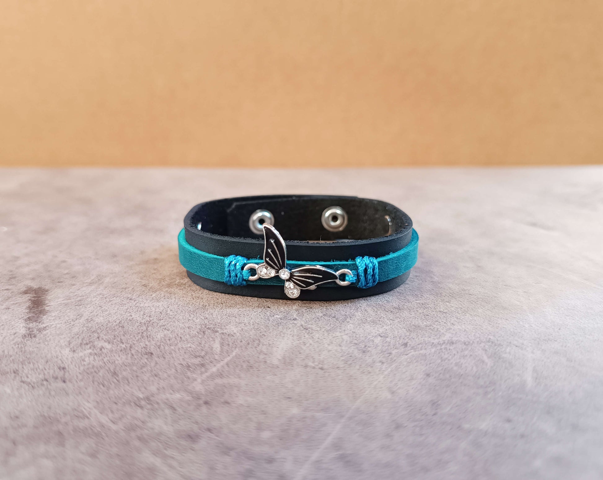 Black leather bracelet with blue cord and black silver butterfly charm on a beige surface
