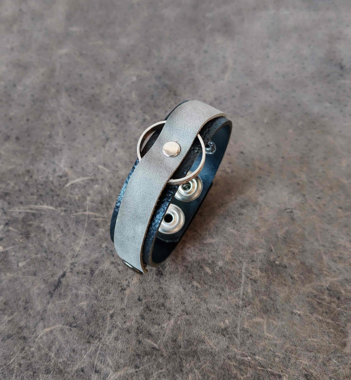 A handmade leather wrap bracelet with a light gray suede-like outer band, a dark blue leather inner band, and silver metal grommets and rings, resting on a textured gray surface.