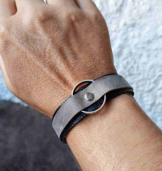A close-up of a person's wrist wearing a dark gray leather wrap bracelet with a silver-tone metal ring and snap closure.