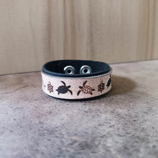 A two-layered leather bracelet with a black base band and a white top layer featuring turtle patterns, with adjustable stainless steel snap buttons.