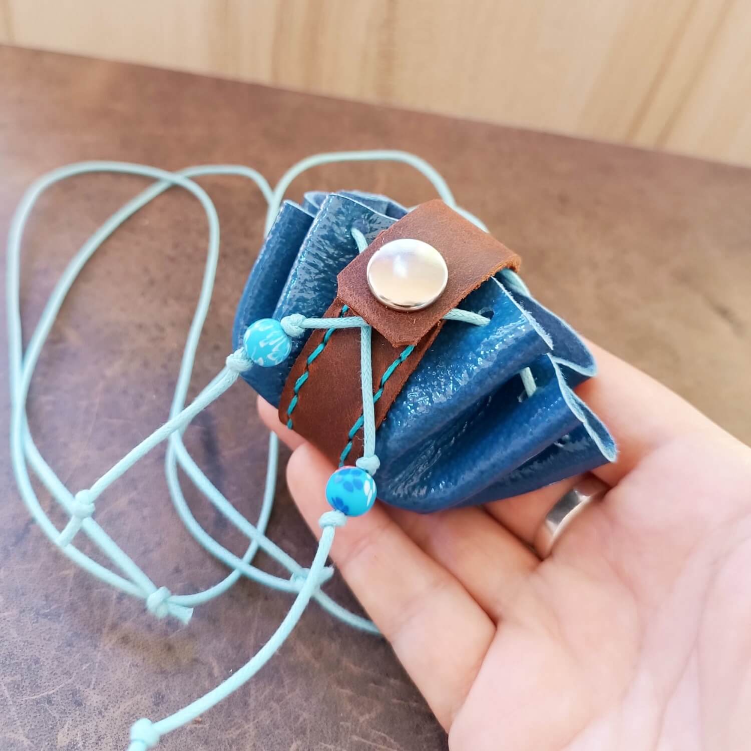 Hand holding a blue and brown mini leather neck pouch with beads, showcasing its tiny size and drawstring closure with the secure snap button.