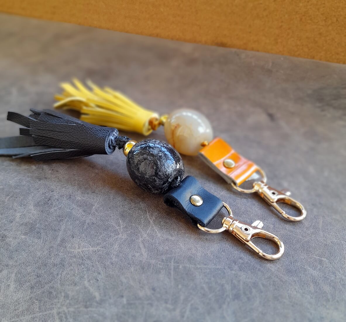 two handmade leather bag charm Keychains with tassels and large beads on a textured surface, one black and one yellow
