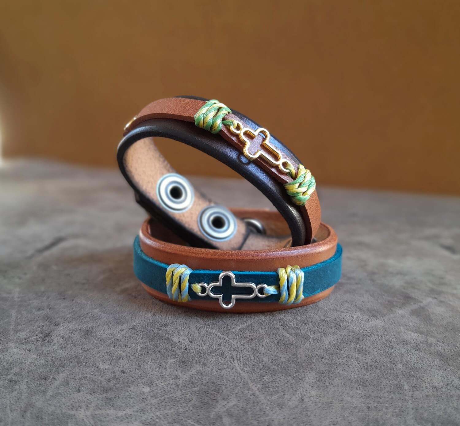 both variations of the cross bracelets, the warm brown with silver cross and blue strap, and the dark brown with gold cross and light brown strap.