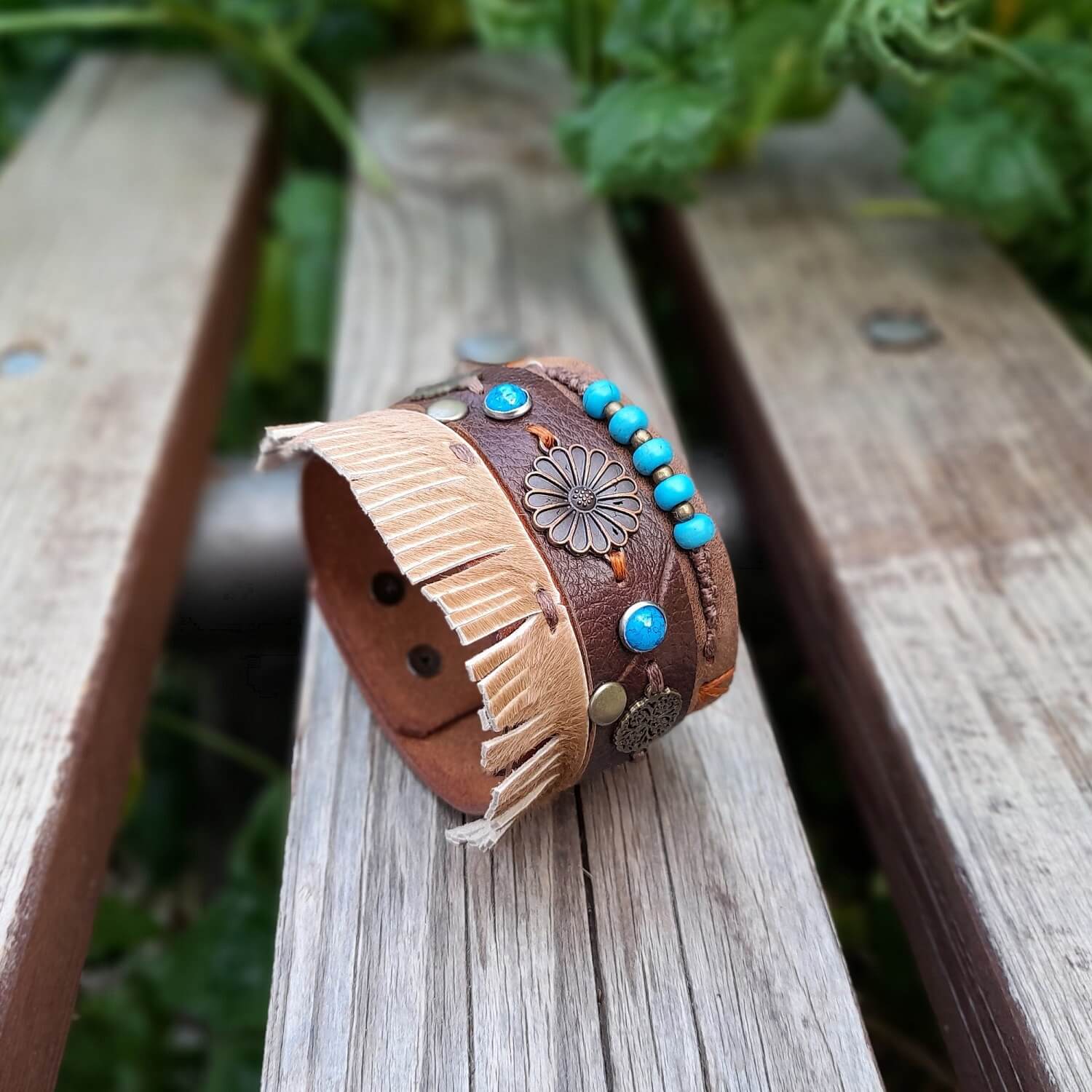 A brown chunky boho leather cuff with floral charms and fringe, displayed on a wooden bench with greenery in the background.