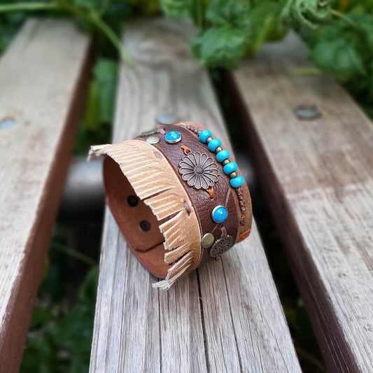 A brown chunky boho leather cuff with floral charms and fringe, displayed on a wooden bench with greenery in the background.