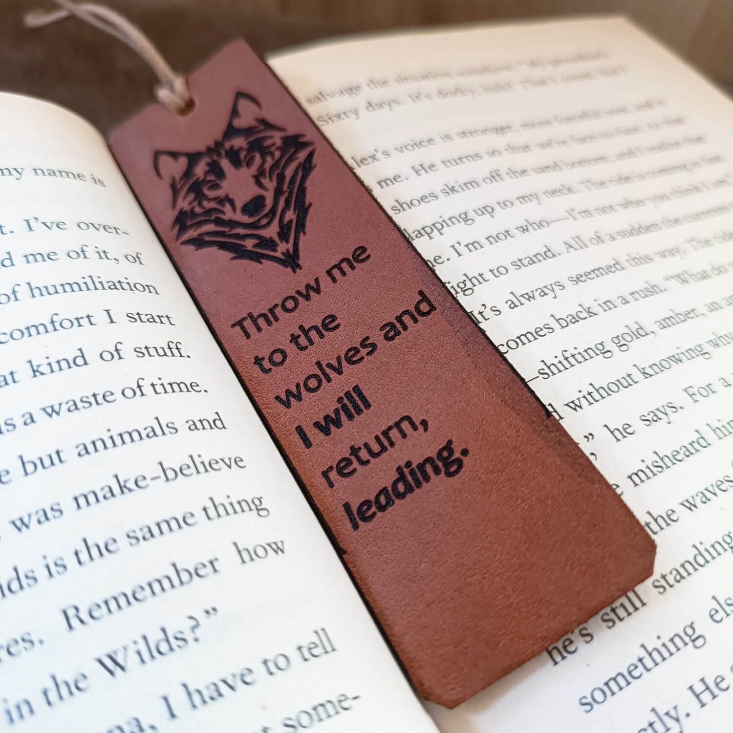A brown leather bookmark with a laser-engraved wolf head and the text 'Throw me to the wolves and I will return, leading.' on it, placed on an open book.