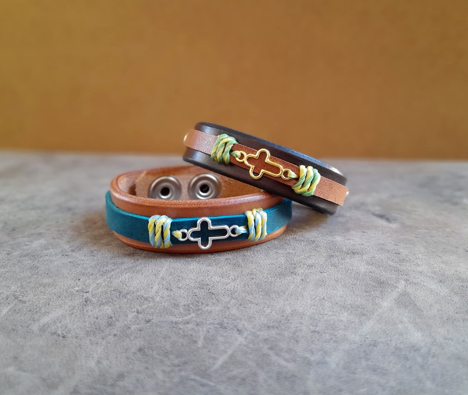 group photo of two brown leather bracelets, one in warm brown and one in dark brown, with silver and gold cross and colorful waxed cords